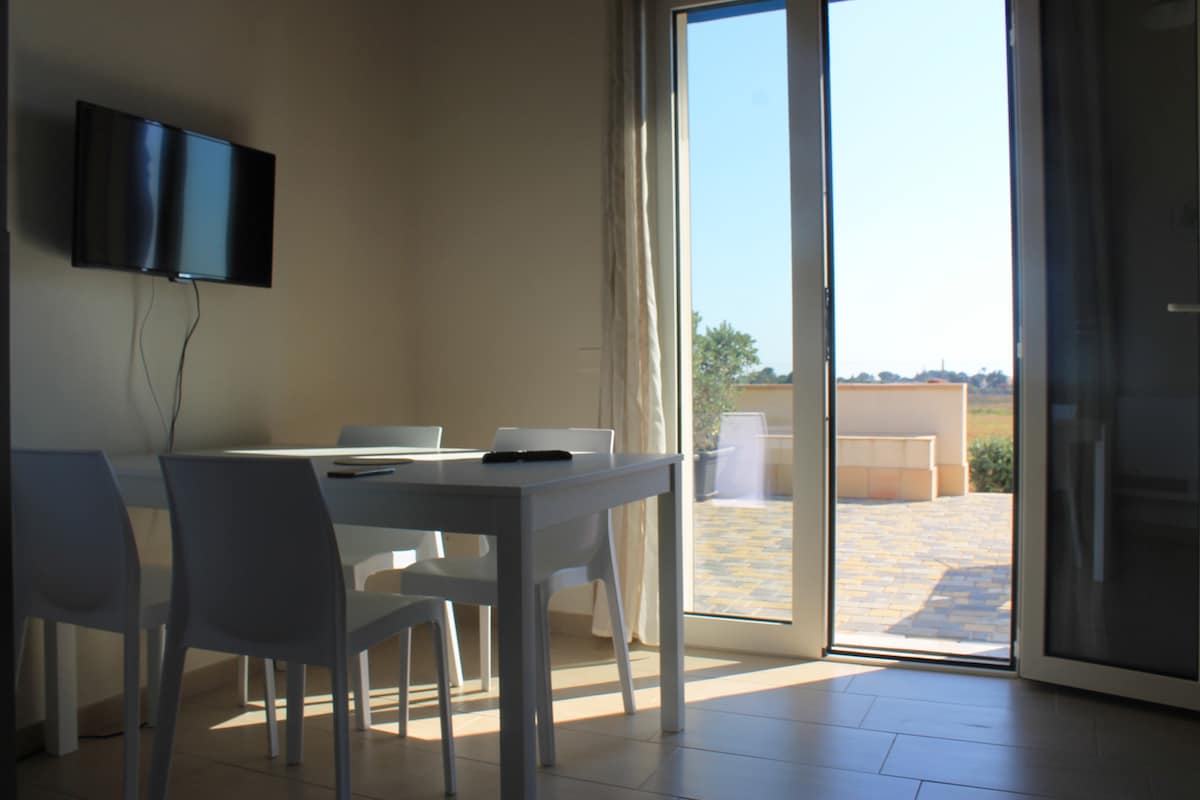 A bright dining area features a white table surrounded by four white chairs. Large glass doors lead to a sunny outdoor terrace, allowing for ample natural light to fill the space.