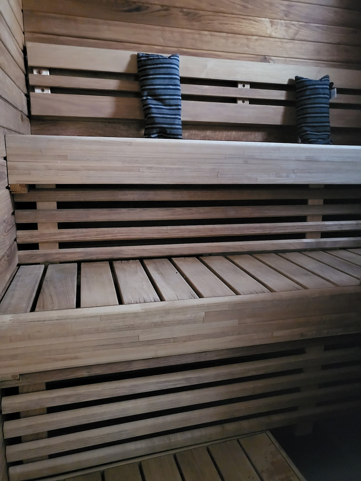 A private sauna features wooden benches arranged at multiple levels, offering a space for relaxation. Two striped cushions are placed on the upper bench, enhancing comfort. The rich texture of the wood creates a serene environment for guests.
