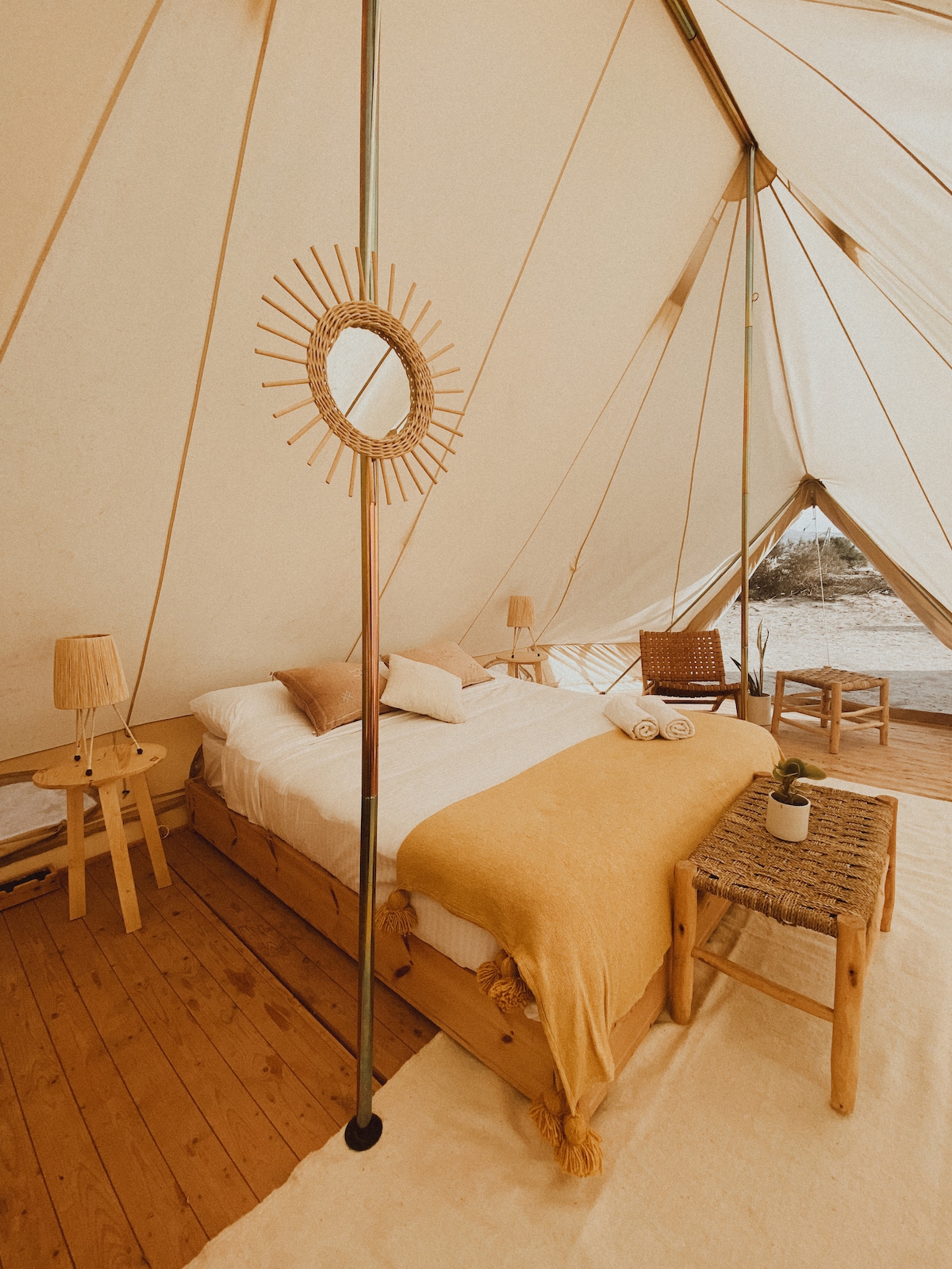 A furnished glamping tent features a spacious bed with soft linens and a light blanket. Natural light filters through the tent, illuminating the wooden floor. A cozy sitting area and decorative mirror add to the inviting space, complemented by a small table and succulent plant.