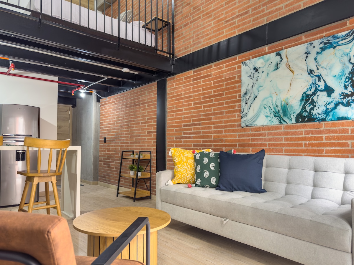 A spacious living area is highlighted by exposed brick walls and modern furnishings. A comfortable sofa with colorful accent pillows faces a round coffee table. The open kitchen is visible in the background, featuring sleek appliances and a bar with seating.