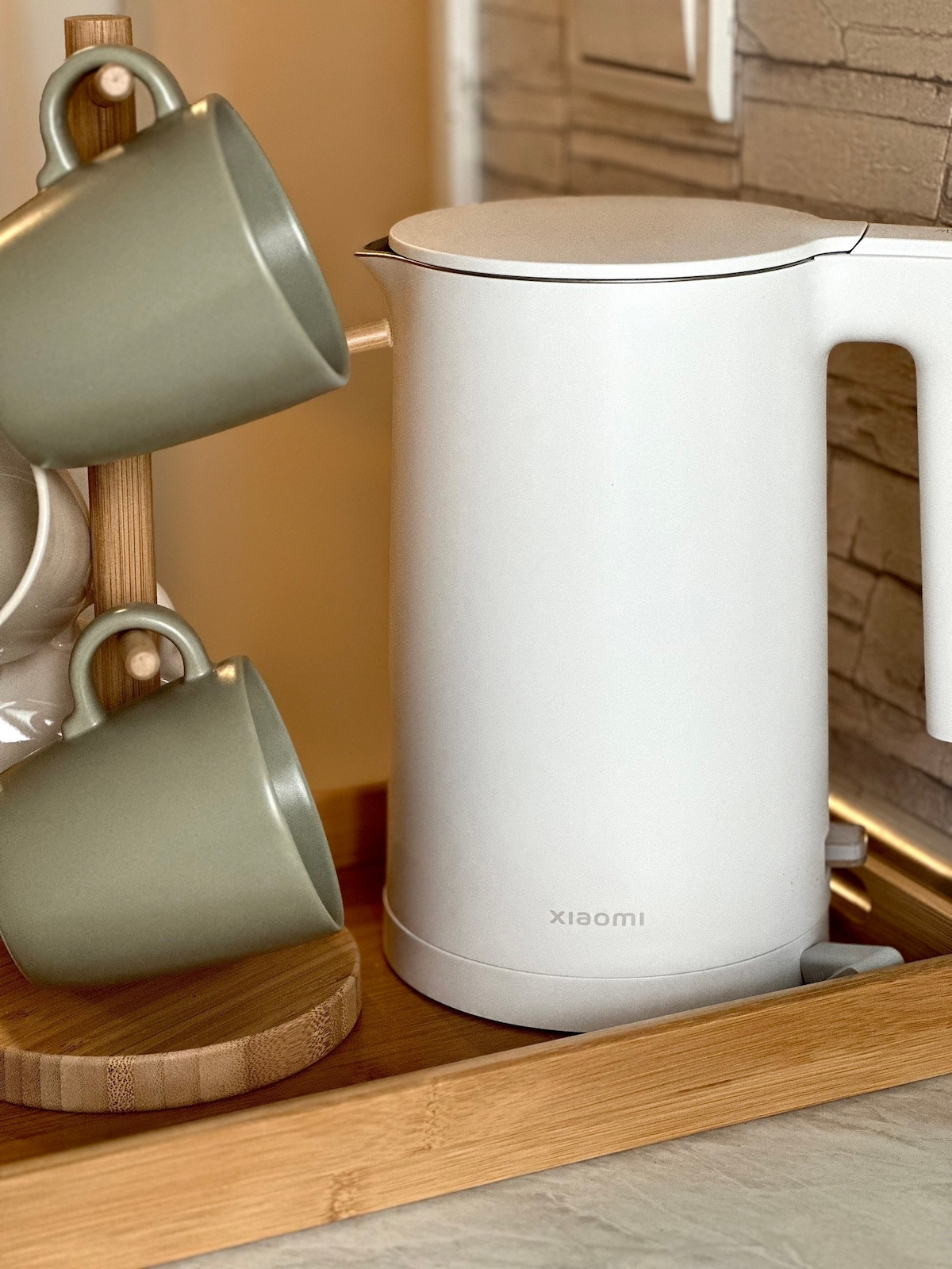 A sleek white electric kettle is positioned next to a wooden tray featuring ceramic mugs. The mugs are displayed on a vertical holder, showcasing a soft green hue that complements the modern design of the kettle.