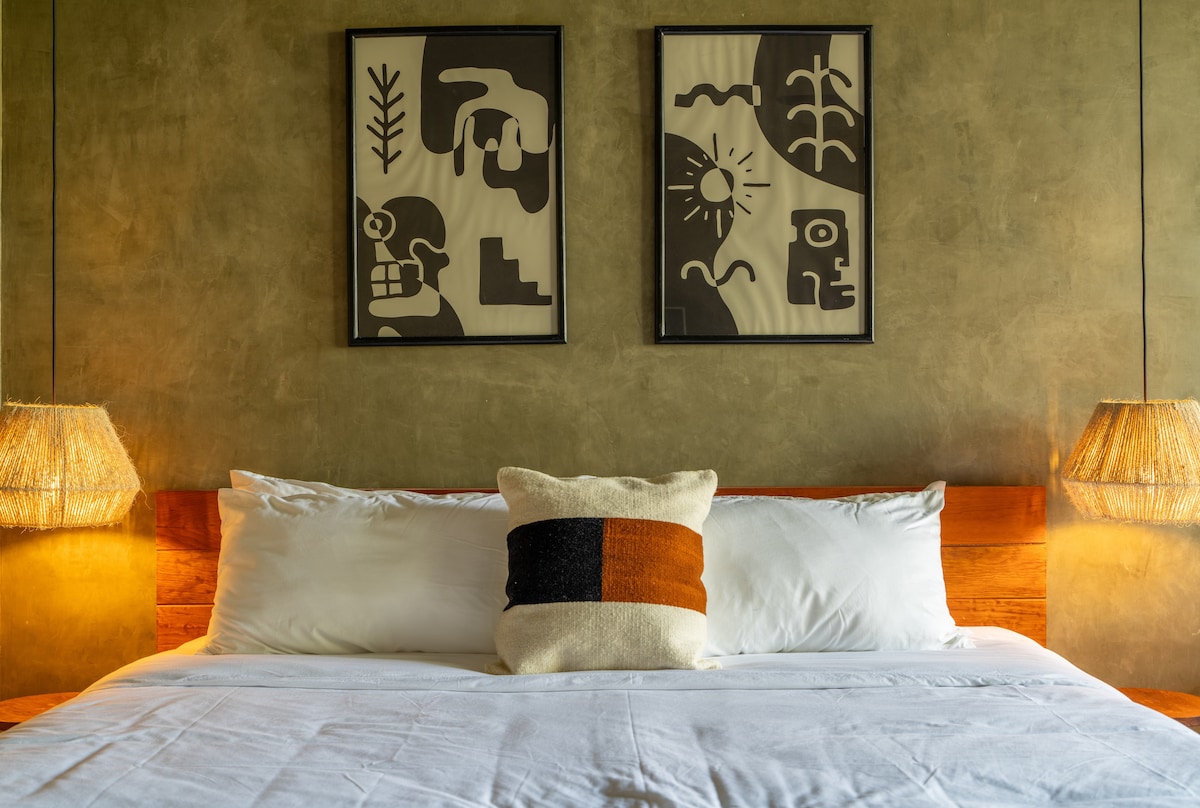 A cozy sleeping area features a spacious bed with crisp white linens. Two decorative wall art pieces are displayed above the bed, showcasing abstract designs. Soft lighting from two bedside lamps creates a warm ambiance against the textured green walls.