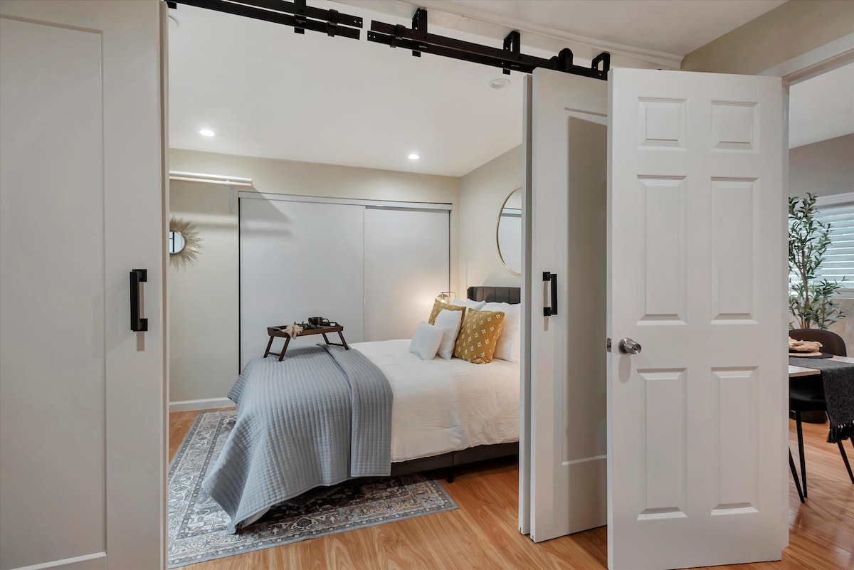 A sliding barn door opens to reveal a cozy sleeping area featuring a spacious bed adorned with decorative pillows. Natural light fills the room, highlighting a small, elegant table positioned at the foot of the bed. A soft area rug enhances the warm wooden flooring.
