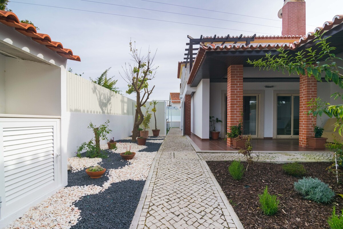 A well-maintained pathway, lined with a variety of potted plants and decorative stones, leads to the entrance of the villa. The exterior features a combination of brick and white siding, while a canopy provides shade over the patio area. Ample natural light brightens the entrance.