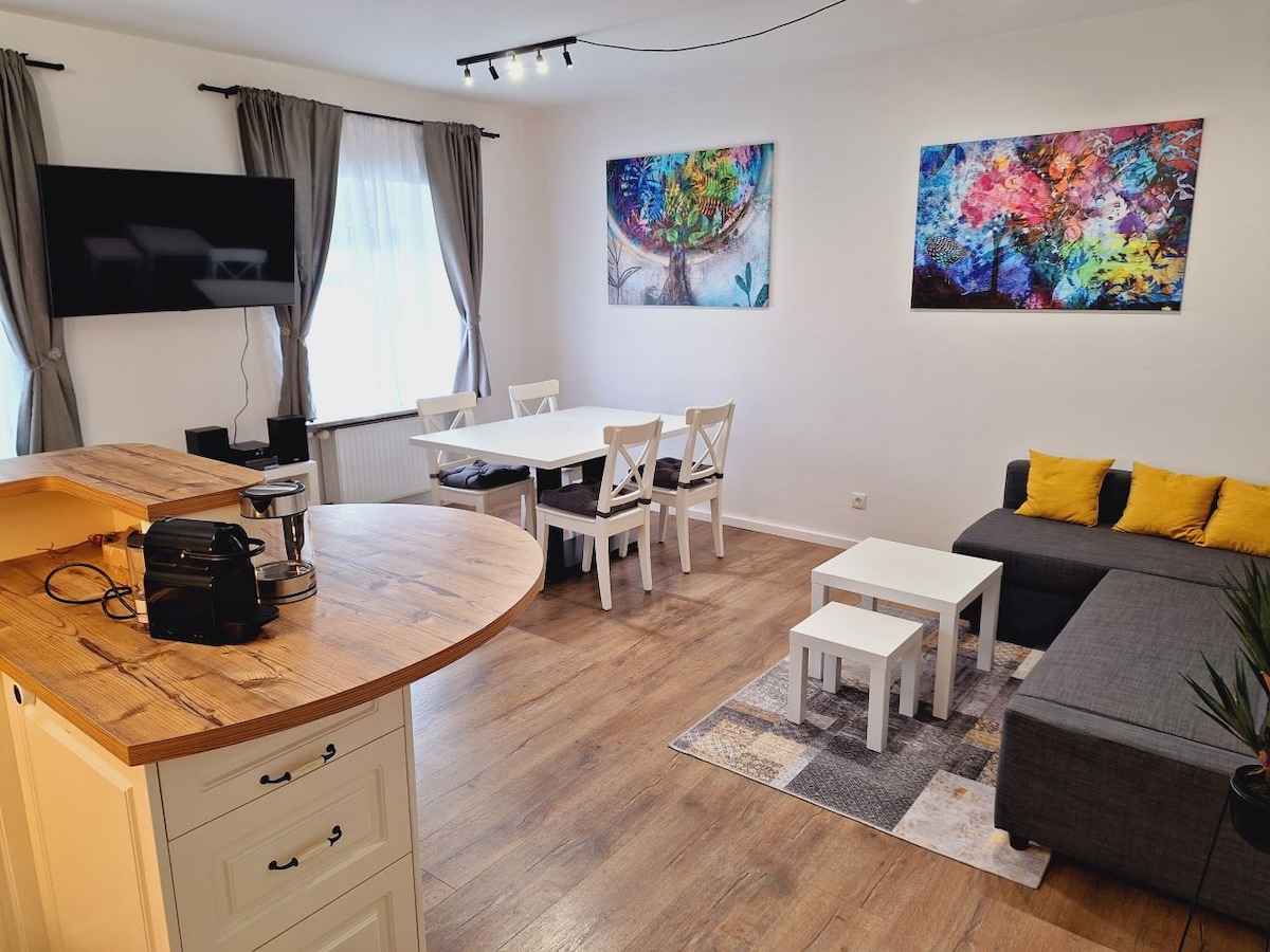 The living and dining area features a modern design with a spacious dining table surrounded by white chairs. A gray sofa sits comfortably in front of a large television, complemented by decorative pillows. Colorful artwork adorns the walls, and light wood flooring enhances the bright ambiance.