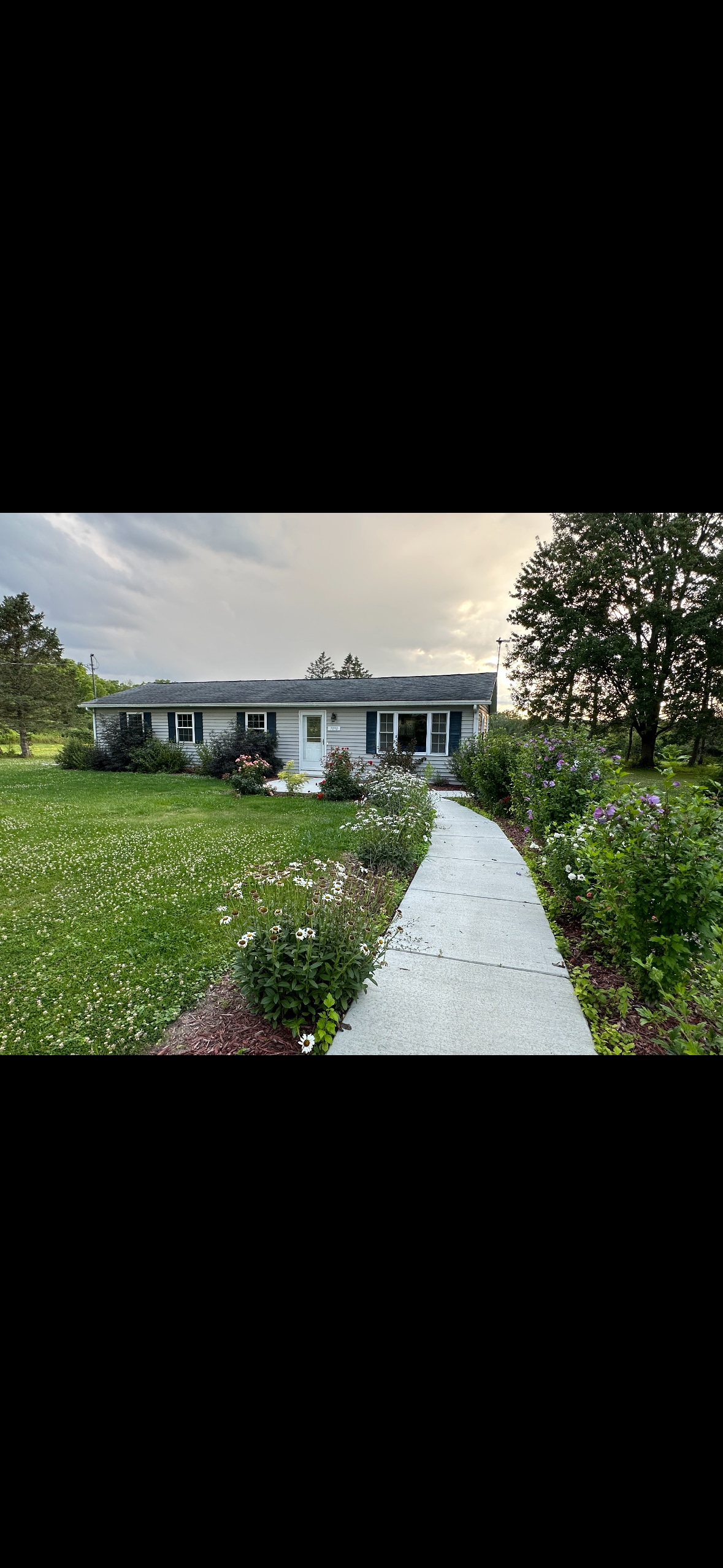 The exterior of the ranch home is surrounded by lush greenery and flower beds, highlighting a peaceful environment. A concrete pathway leads to the front door, with the house displaying large windows that invite natural light.