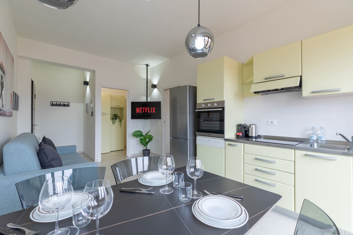 A modern kitchen area is showcased with sleek yellow cabinetry and stainless steel appliances. A dining table is set for four, with clear glassware and plates arranged neatly. A cozy sofa is visible in the background, accompanied by a wall-mounted screen displaying a streaming platform.