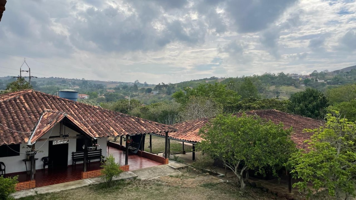 A peaceful landscape showcases two buildings with terracotta roofs surrounded by greenery. Rolling hills are visible in the distance under a sky with scattered clouds. A porch area is positioned in front of the main house, offering tranquil outdoor views.