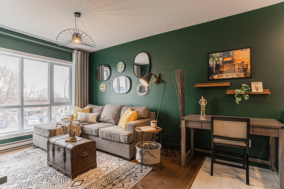 A cozy living area features a gray sectional sofa adorned with decorative pillows. A mix of round mirrors decorates the dark green wall, while a small workspace with a wooden desk and chair is positioned beside it. Natural light floods in through the large windows.