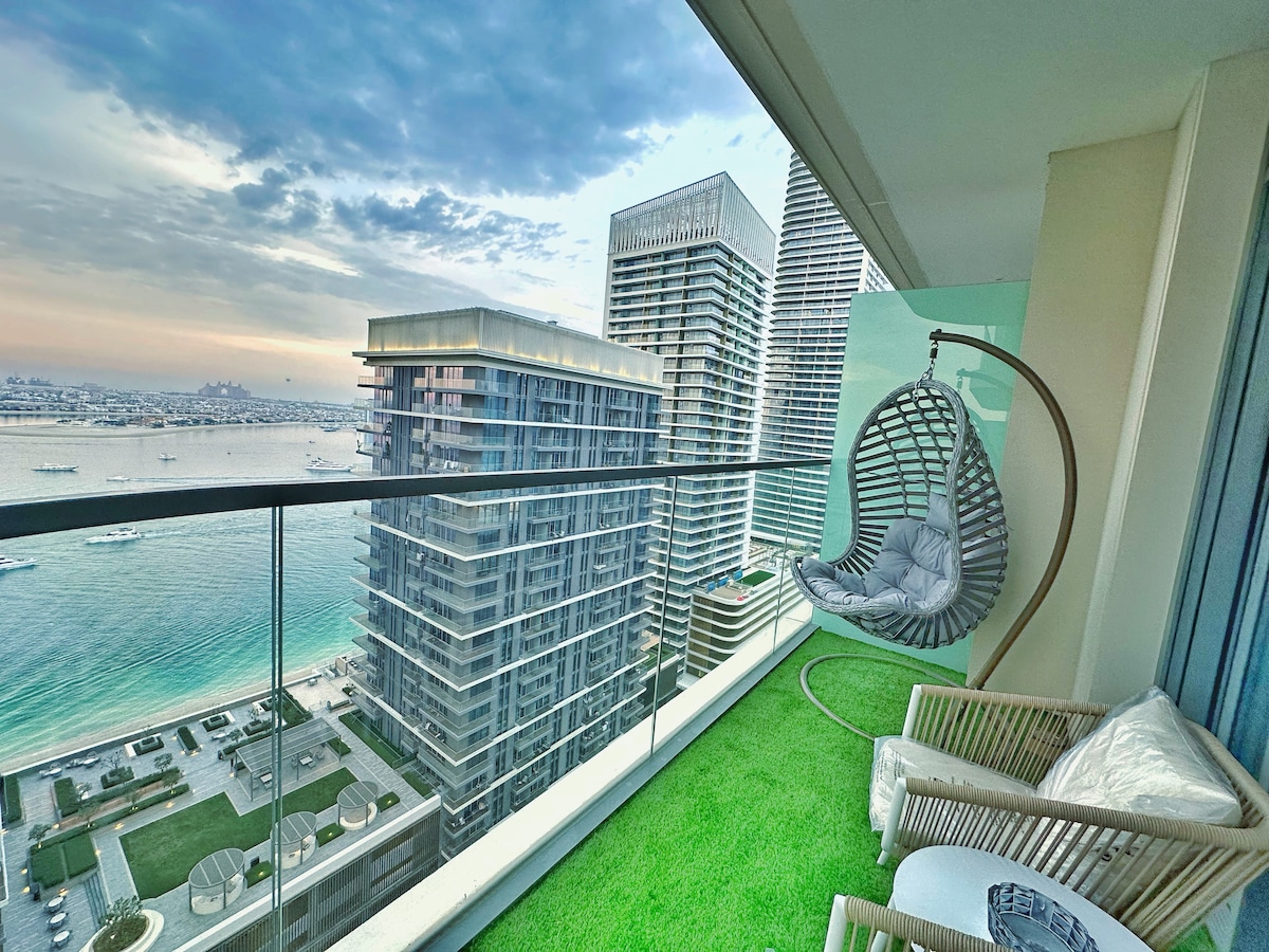 A balcony space features a hanging chair and a comfortable seating area. Lush green grass covers the balcony floor. The view overlooks a serene waterfront with boats, and modern buildings rise against a cloudy sky.