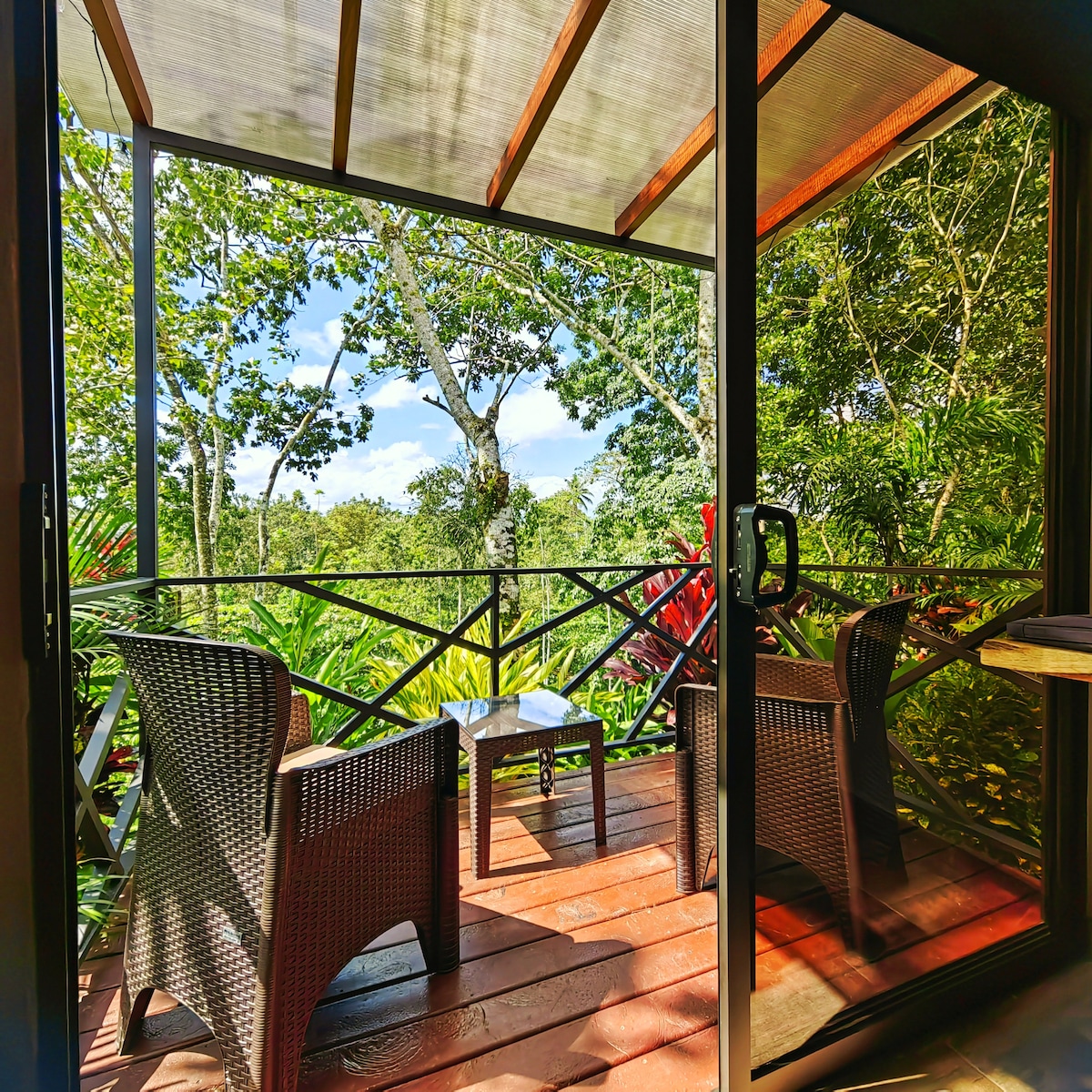 A screened balcony features two dark wicker chairs with a small table between them. Lush greenery is visible through the glass doors, with sunlight filtering through the leaves, creating a serene outdoor space perfect for relaxation. The wooden floor adds warmth to the setting.
