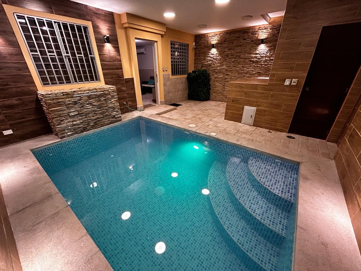An indoor pool area is showcased, featuring clear blue water with steps leading into it. Soft lighting illuminates the space, highlighting textured stone walls and a large window. The tranquil atmosphere is complemented by plants placed strategically around the pool.