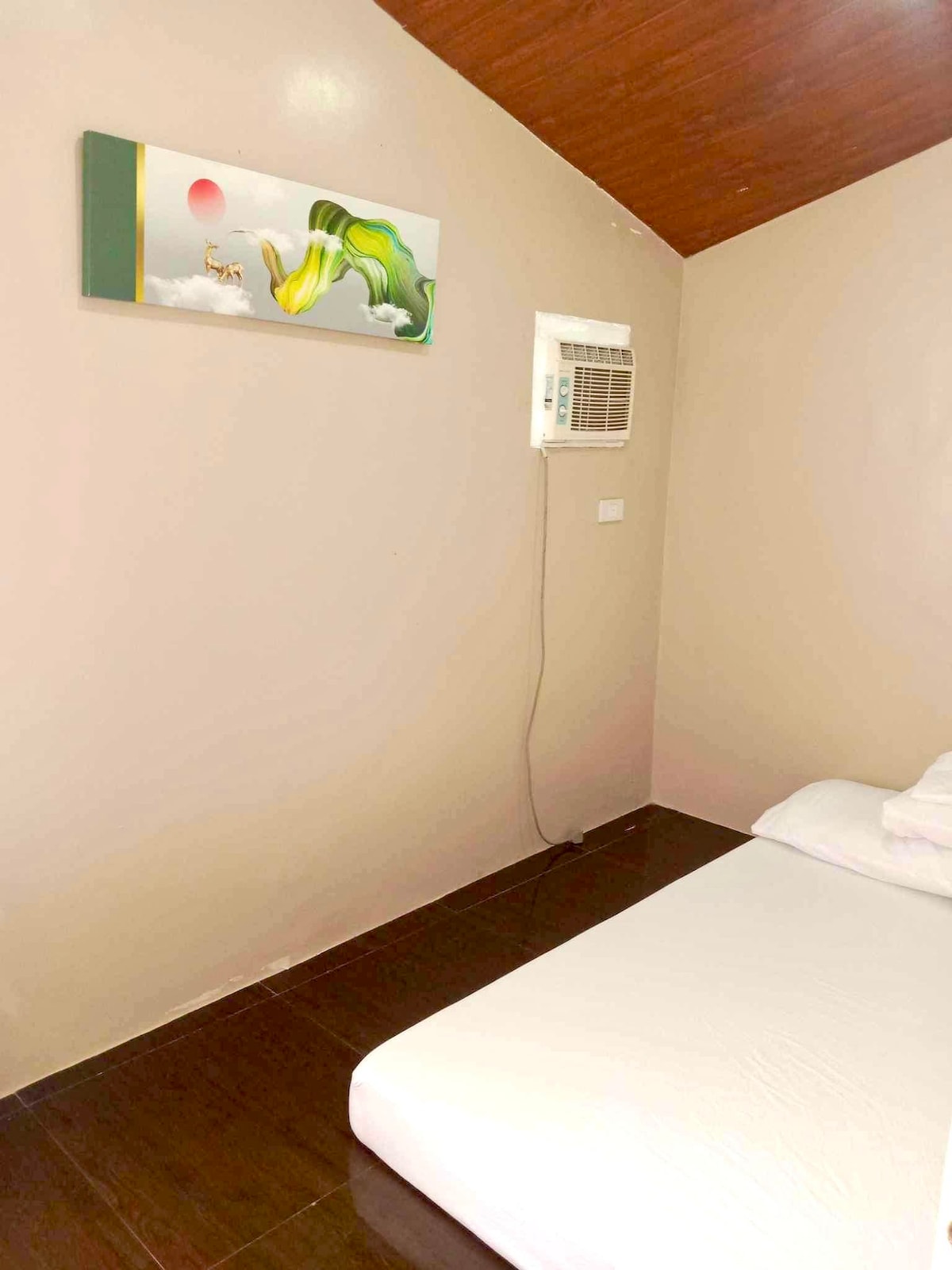 A minimalist sleeping area is featured with a white mattress positioned against a light-colored wall. An abstract artwork hangs above, contrasting with the dark wood flooring. An air conditioning unit is mounted on the wall, providing comfort to the space.