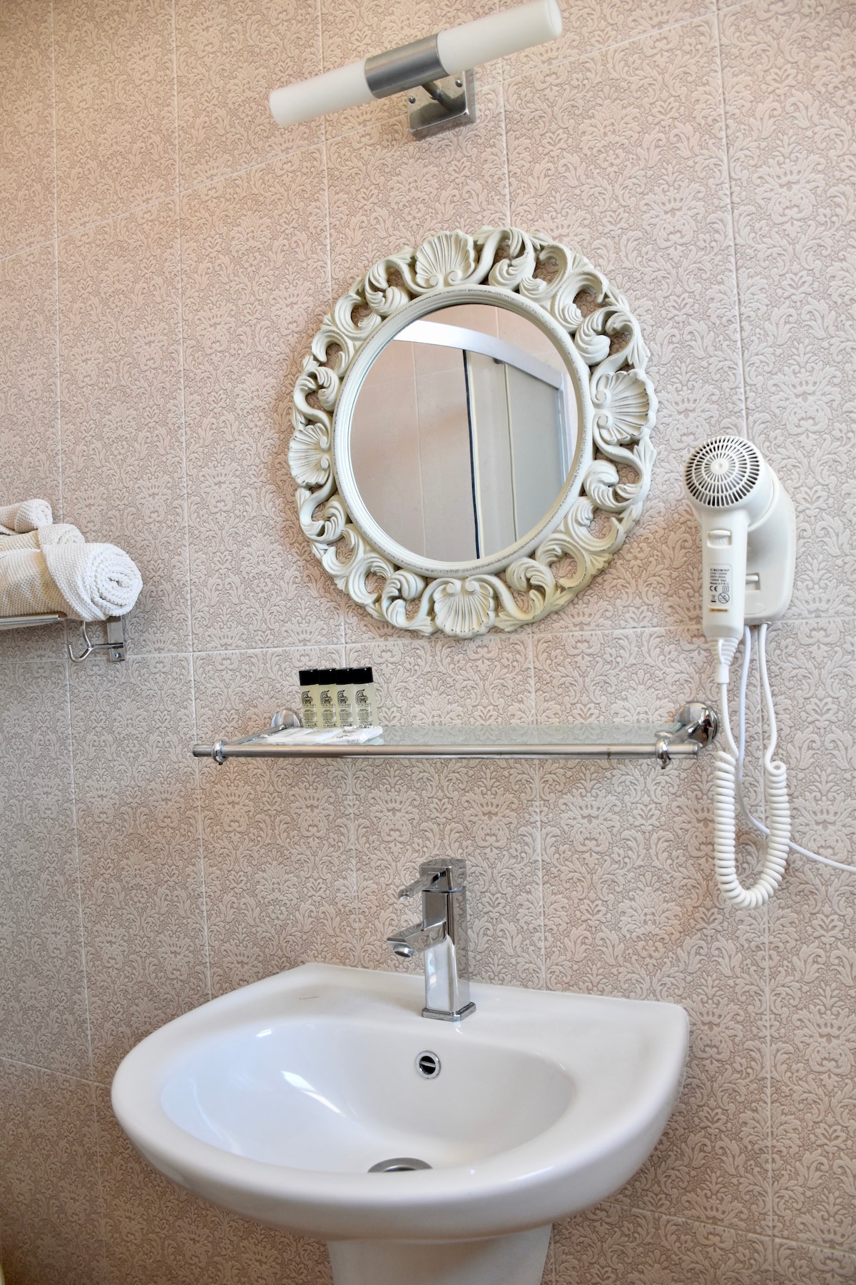 A compact bathroom space is highlighted by a decorative mirror with a scalloped frame. A small sink with a chrome faucet is positioned below the mirror. Fresh towels are neatly arranged on a rack, and toiletries are displayed on the shelf.