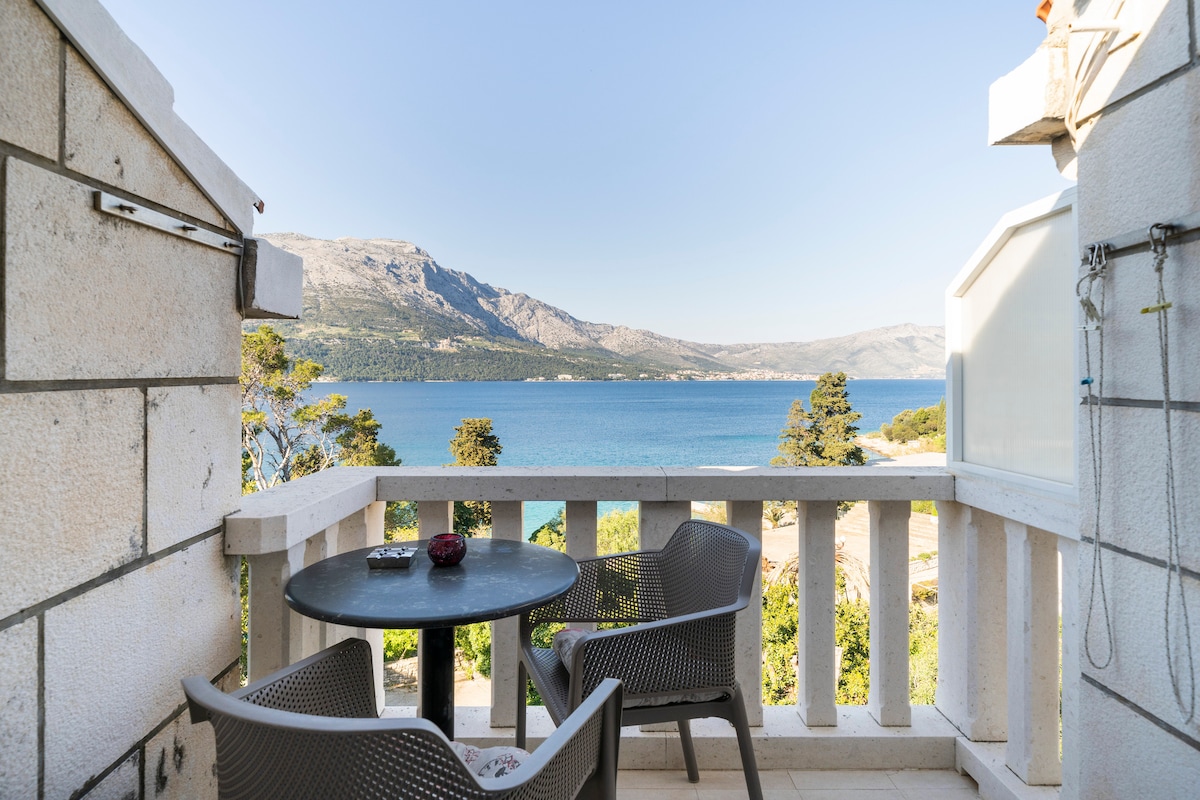 A terrace features a round table with two chairs, offering a clear view of the sea and surrounding mountains. The railing provides a sense of openness, while the natural landscape adds to the tranquil atmosphere of the space.