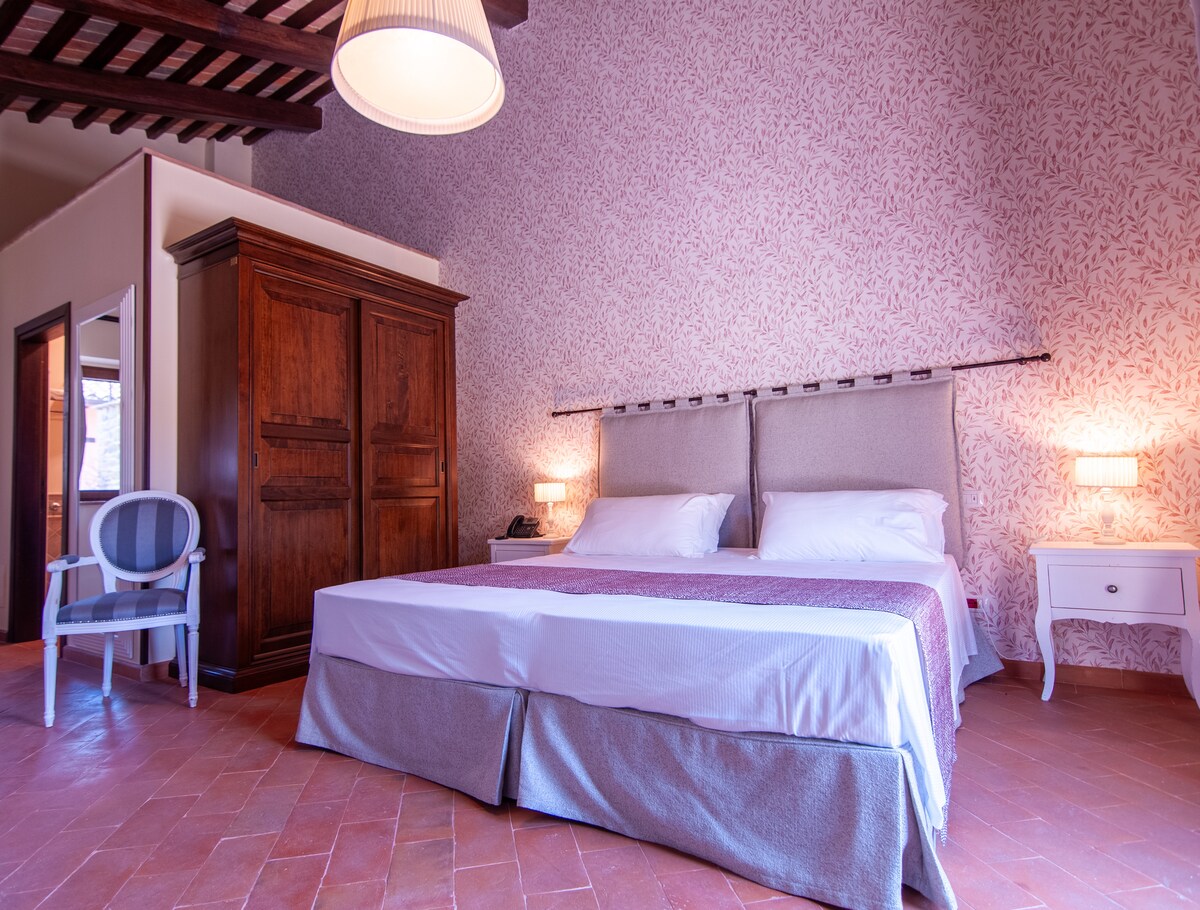 A spacious bedroom features a large bed with soft linens and a decorative headboard. Light wood bedside tables with lamps sit on either side. A vintage wardrobe stands against a floral-patterned wall, while a single chair complements the room's elegant design.