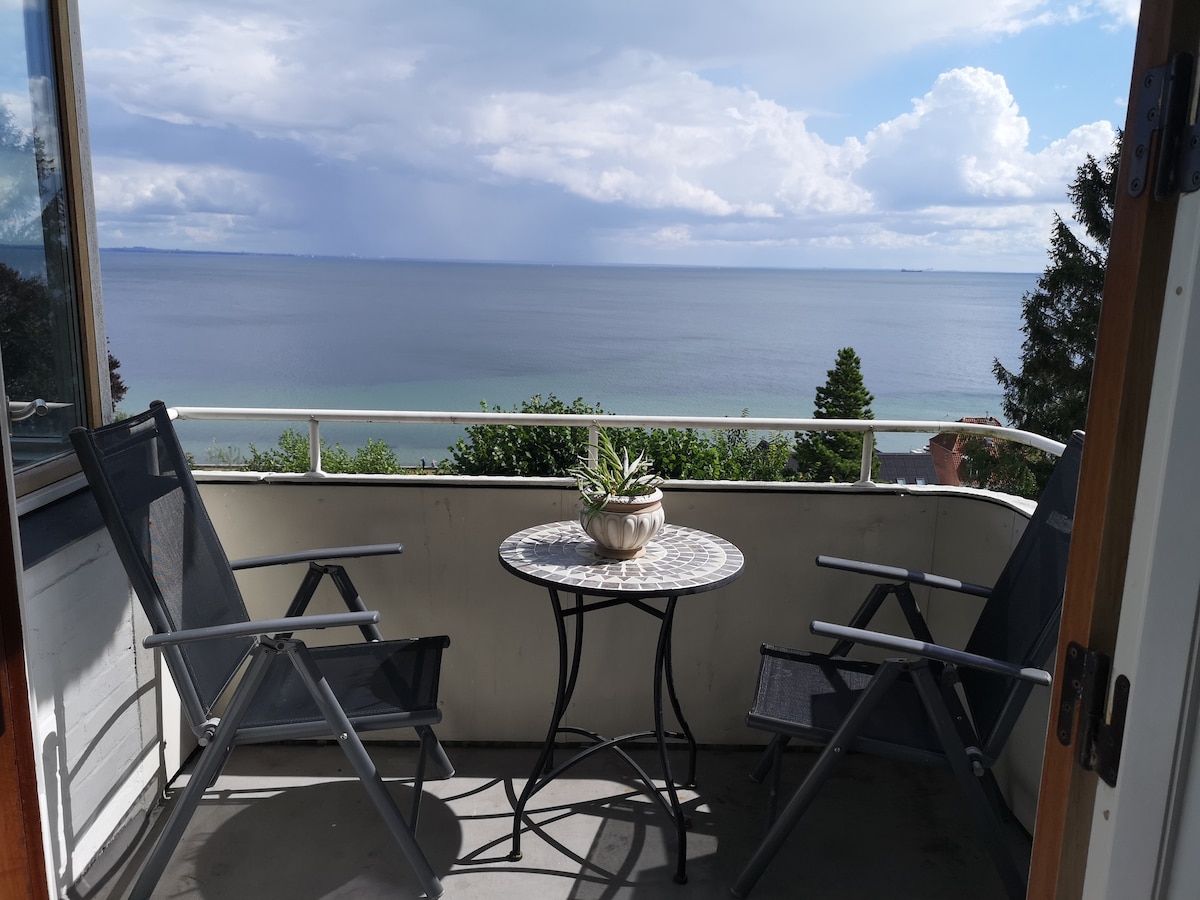 A serene balcony is presented, featuring a small round table topped with a decorative bowl and two reclining chairs. The expansive view of the calm water extends toward the horizon, with trees lining the space, under a partly cloudy sky.