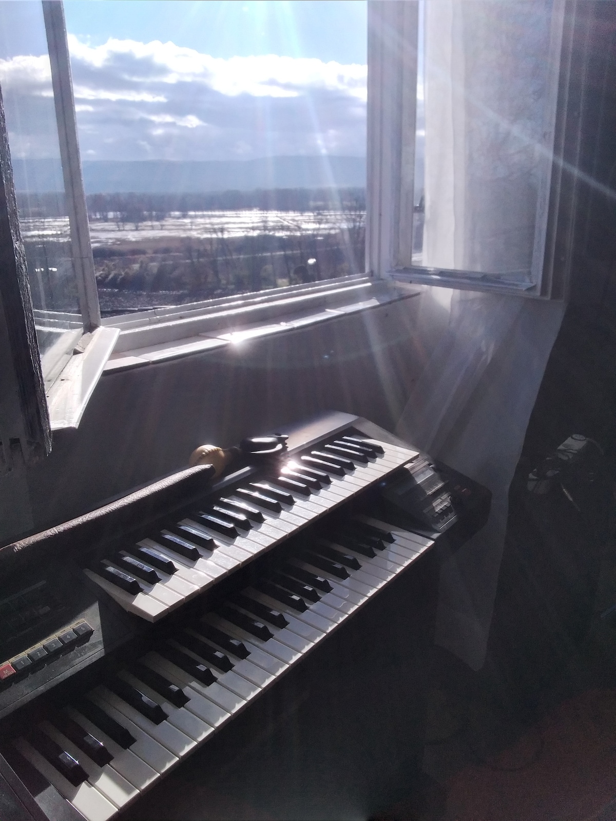A vintage keyboard sits on a table, with sunlight streaming in through an open window, creating a bright atmosphere. The window offers a view of a landscape filled with distant trees and shimmering water, enhancing the space's natural light.