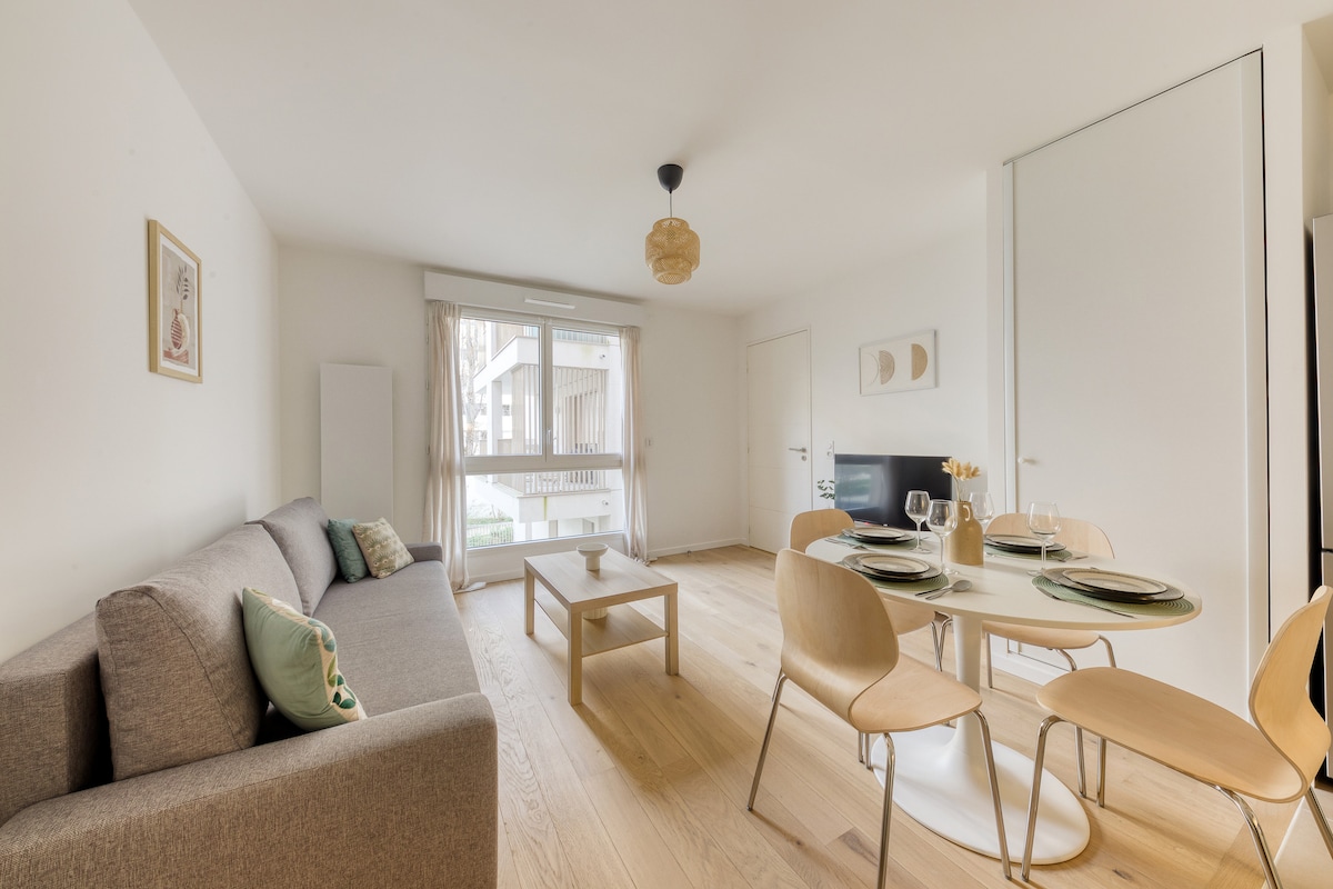 A modern living area is displayed, featuring a gray sofa and a light wood coffee table. A dining area with a round table and four chairs is positioned nearby. Natural light fills the room through large windows with sheer curtains, enhancing the open layout.