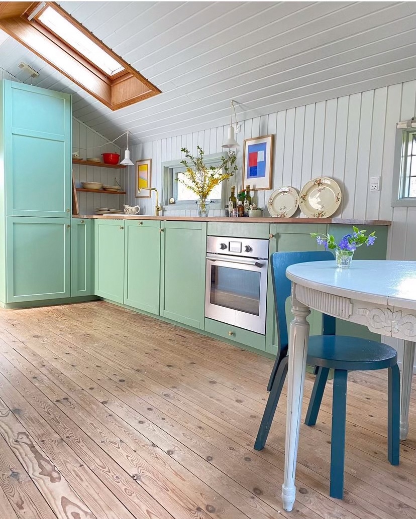 A bright kitchen is featured with light green cabinetry and wooden countertops. A built-in oven and shelves hold kitchen items. Natural light filters in through a skylight, illuminating the wooden floor. A small round table with a blue chair and a vase of flowers adds a touch of color.