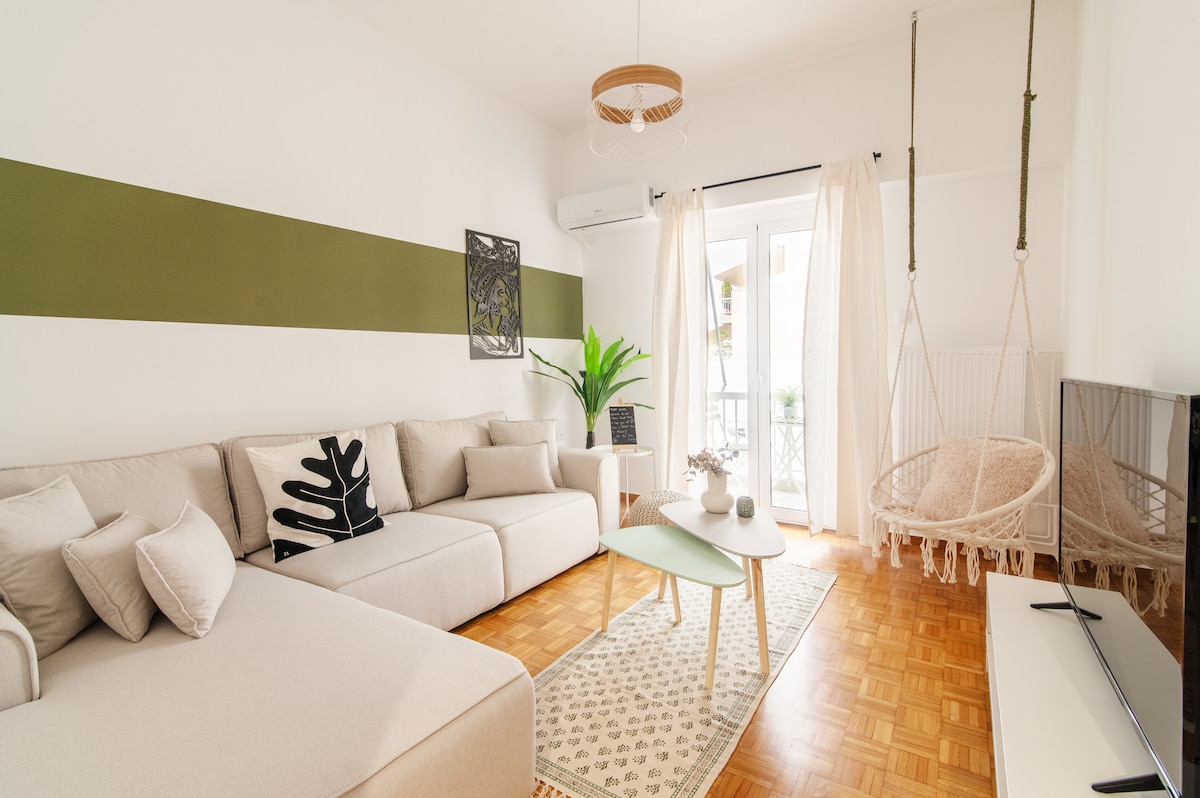 A bright living area is presented with a spacious light-colored sectional sofa and a pale green accent wall. A minimalist coffee table sits on a patterned rug, while two hanging chairs add a relaxed vibe. Natural light enters through the balcony doors, with greenery visible outside.