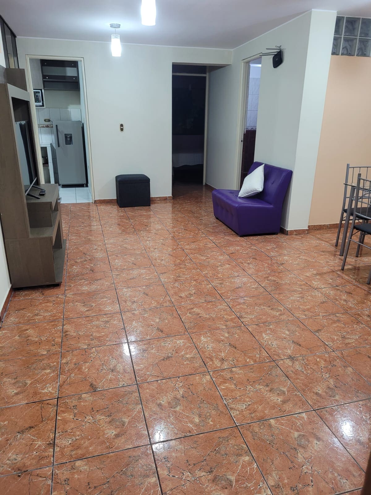 A spacious living area features polished tile flooring and neutral-colored walls. A purple armchair is placed beside a small ottoman, while a dining table with metal chairs occupies one corner. Soft lighting is provided by overhead fixtures, enhancing the room’s open ambiance.