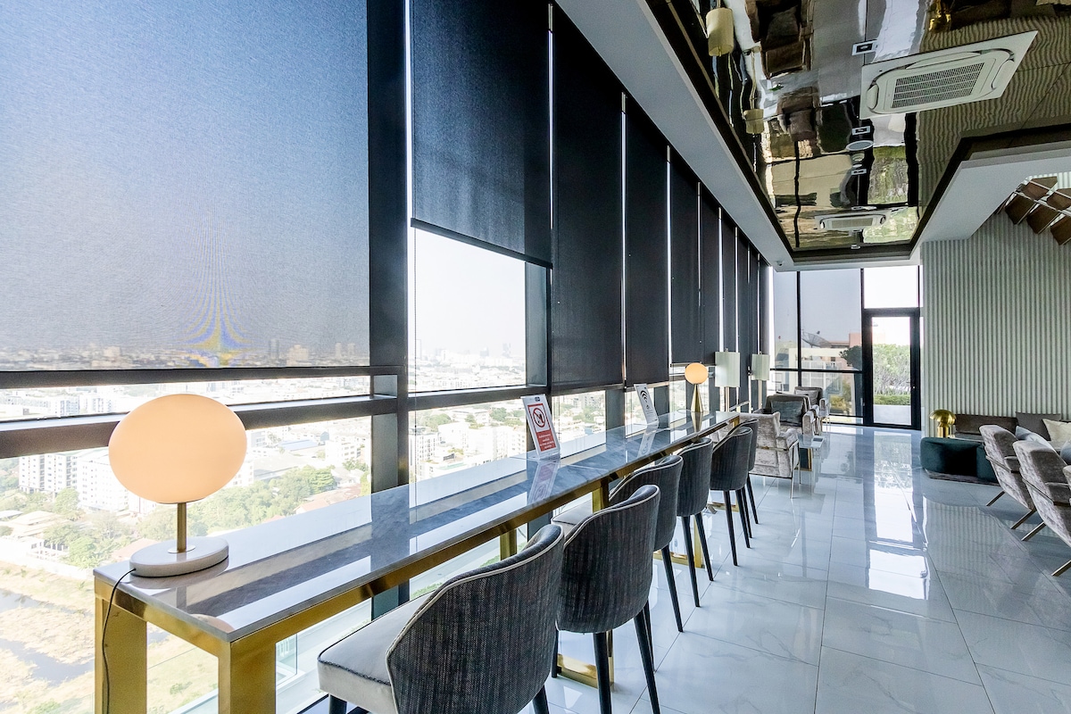 A sleek co-working space is depicted with a row of elegant chairs positioned at a long table under large windows. Natural light filters through the modern blinds, creating a bright atmosphere. A stylish lamp stands on the table, providing additional lighting.