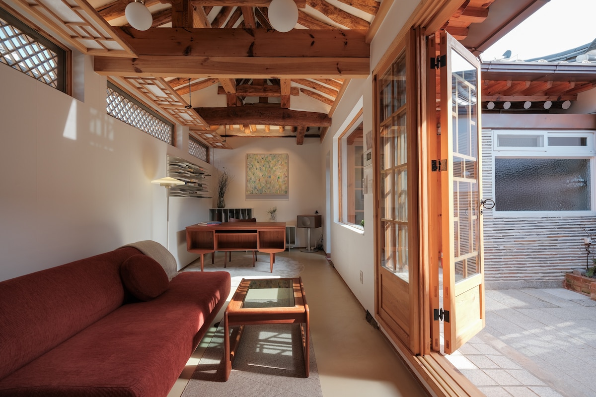 The living area features a brick patio that leads to a wooden-framed glass door, opening to a sunlit space. A red sofa is positioned next to a wooden coffee table, with a modern desk and shelves in the background. Natural wood beams enhance the ceiling design.