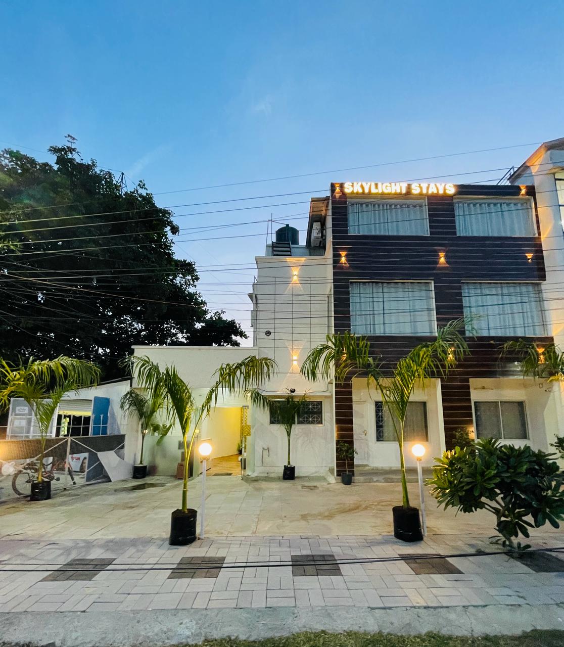 The exterior of Skylight Stays is showcased, featuring a modern building illuminated by stylish lighting. Lush palm trees are positioned beside the entrance. The pathway is framed by square stone tiles, providing an inviting approach.
