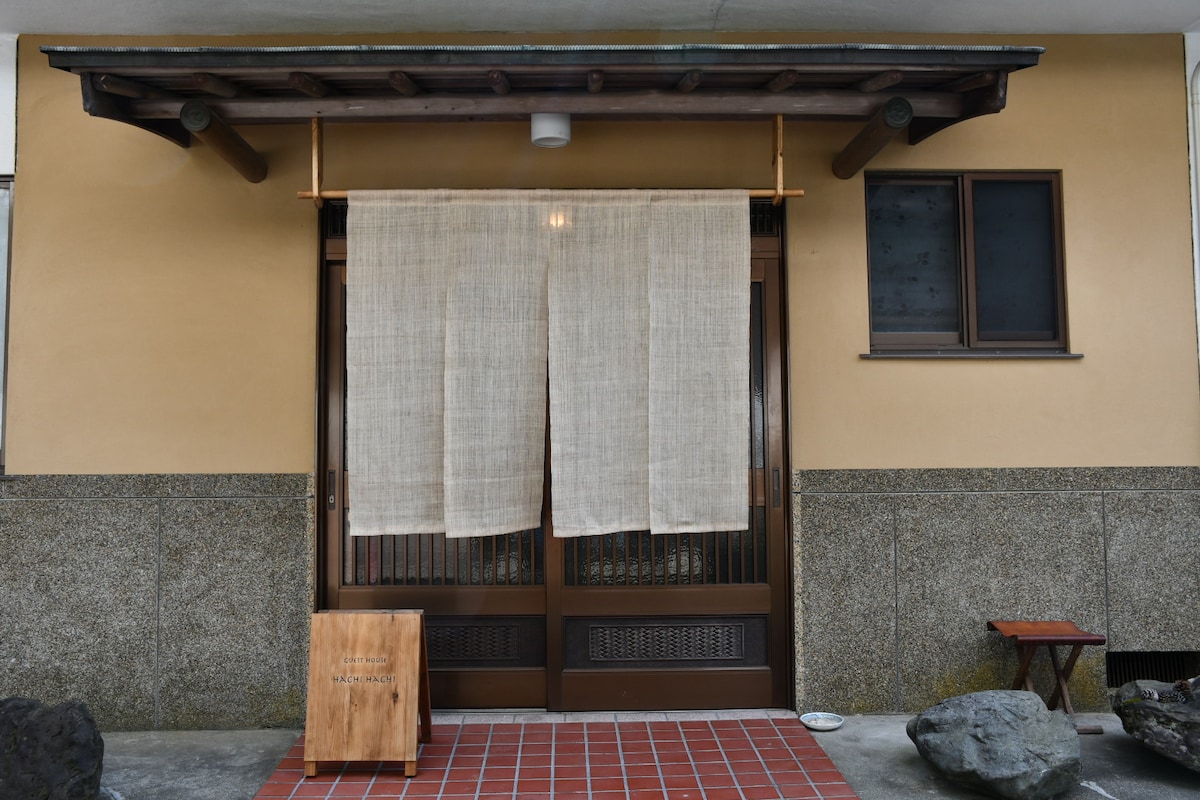 The entrance features a wooden door adorned with a natural fabric curtain, softly draping down. A wooden sign rests beside the door, and a small table with a bench is positioned nearby. The exterior walls are a warm, neutral tone complemented by stone details.