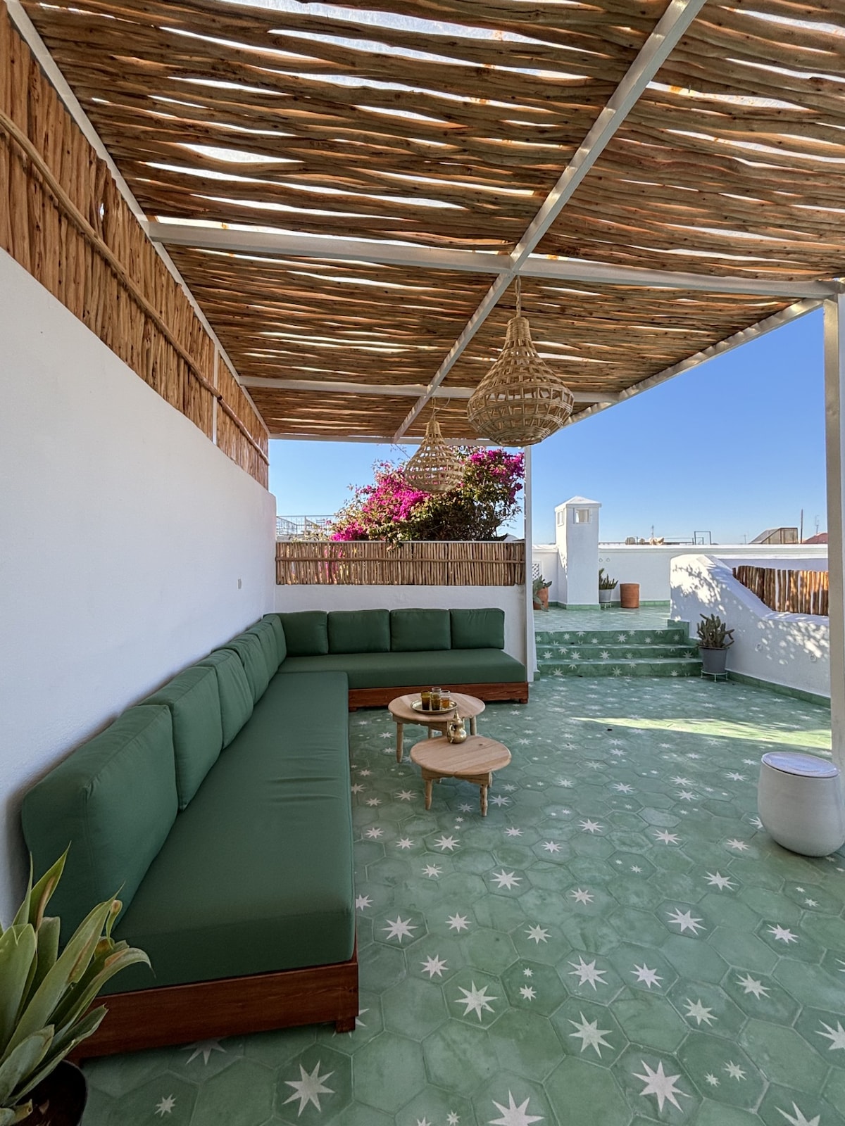 A spacious terrace is adorned with light green tiles and a shaded seating area featuring a large corner sofa. Small wooden tables are placed alongside plants. Vibrant flowers bring color to the surrounding area, while clear blue skies are visible overhead.