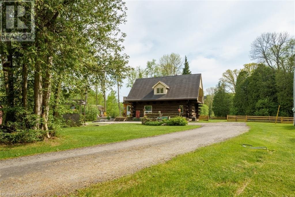 A rustic cabin is set amidst lush greenery, featuring a sloped roof and wooden log exterior. A gravel driveway leads up to the entrance, while trees surround the property, providing a sense of seclusion in a serene natural environment.