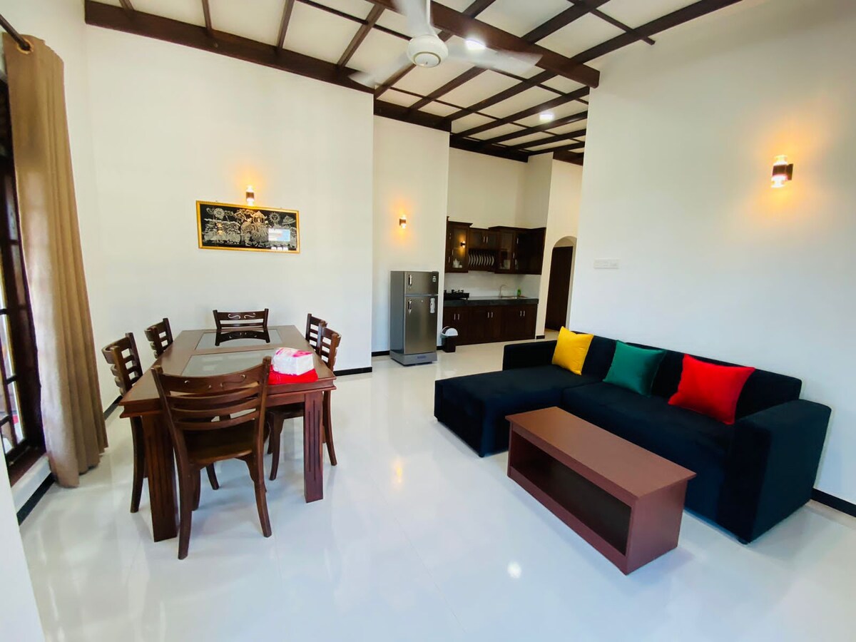 A bright living area features a dark blue sofa with colorful cushions. A wooden dining table with matching chairs is positioned nearby. The space is complemented by a kitchenette in the background, with modern appliances and warm lighting creating a welcoming environment.