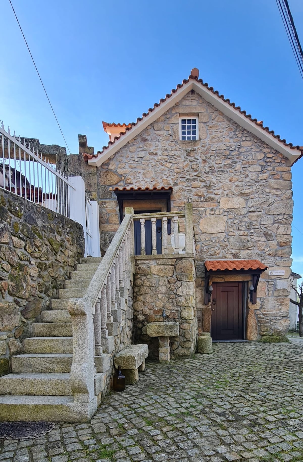 A charming stone house is depicted, featuring a sloped roof with terracotta tiles. A staircase made of stone leads to the main entrance, framed by a wooden door. The surrounding area includes cobblestone paths and natural stone walls, enhancing the structure's rustic appeal.