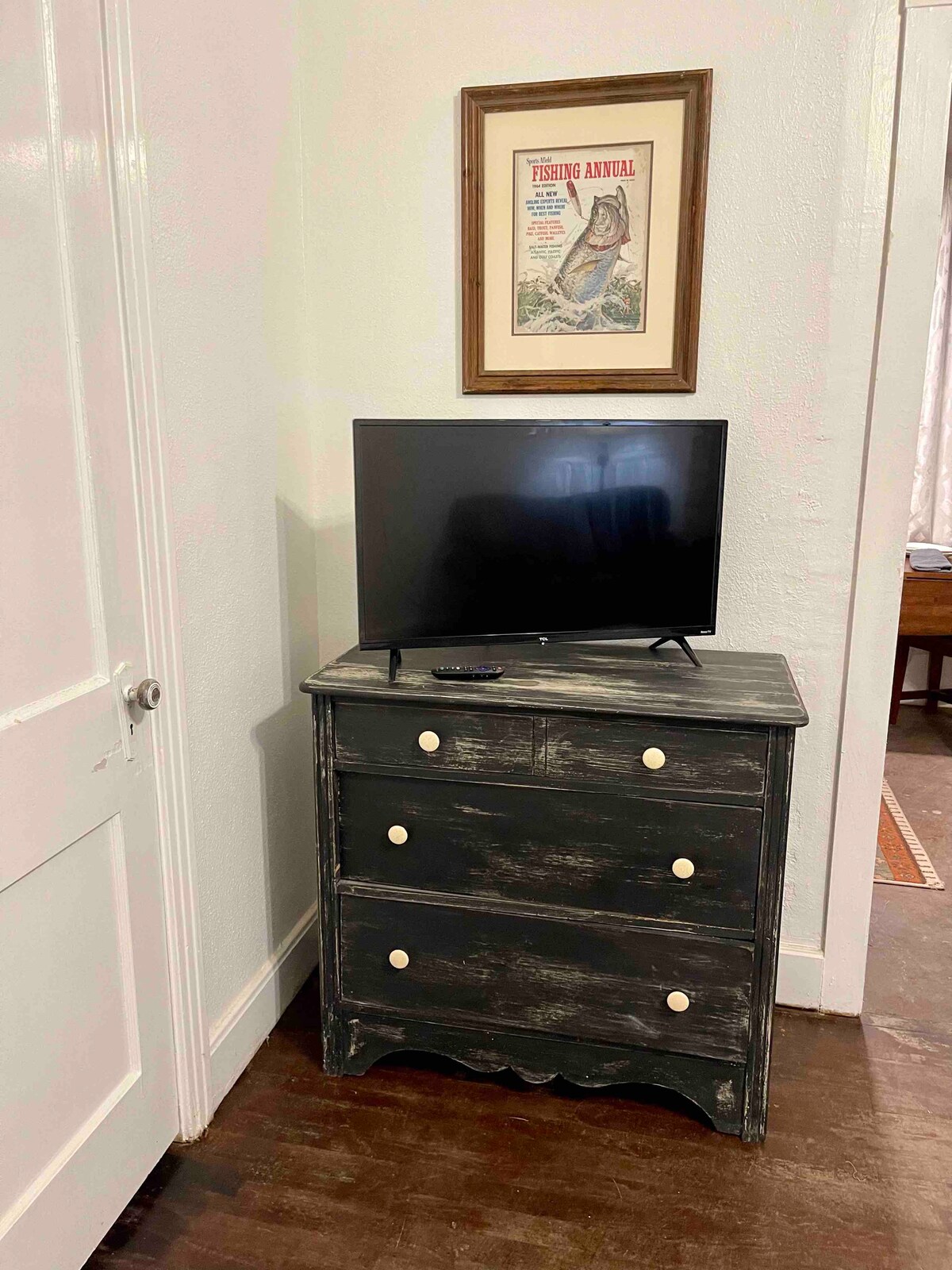 A chest of drawers with three spacious drawers is positioned against a white wall. On top of the chest, a flat-screen television is displayed. Above the furniture, a framed fishing-themed poster adds character to the space.