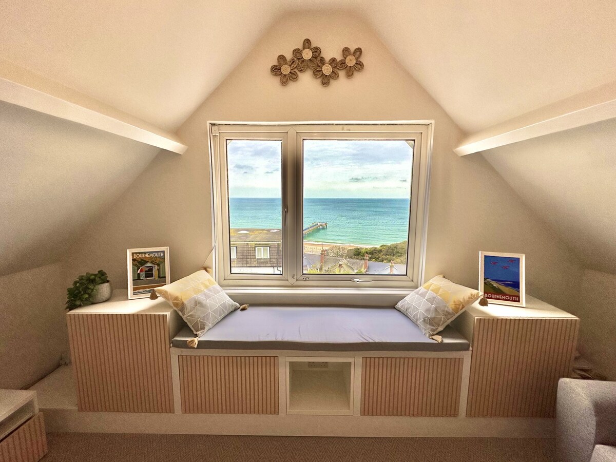 A bright corner nook is featured with a comfortable window seat positioned below two large windows, offering expansive views of the coast. Decorative pillows are arranged, and framed artworks stand on either side, enhancing the cozy ambiance of the space.