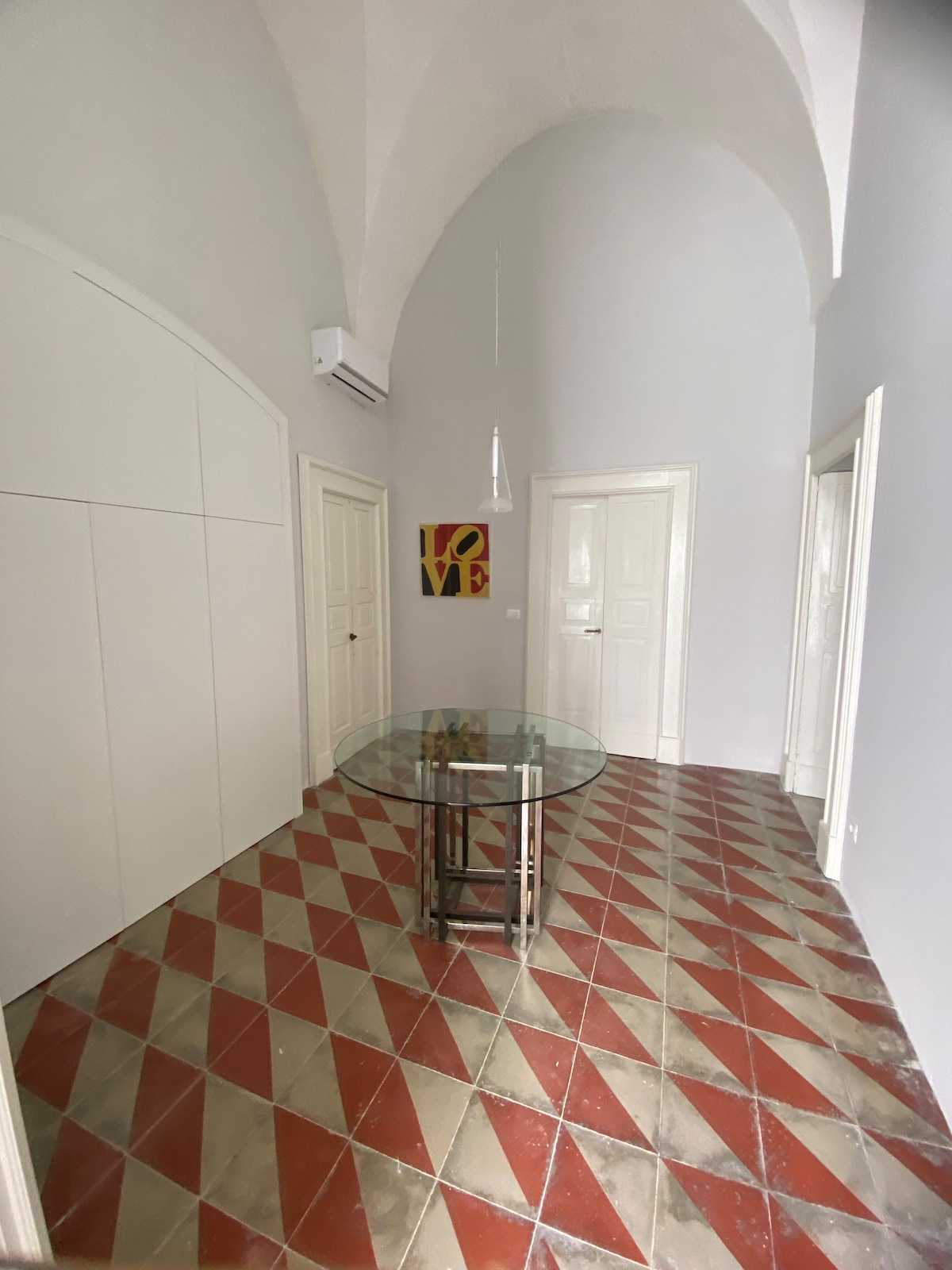 A spacious entryway features a glass table at its center, surrounded by a patterned tile floor with red and cream diamonds. Arched ceilings enhance the sense of space, while two white doors lead to adjacent rooms. A small piece of artwork is visible on the wall.