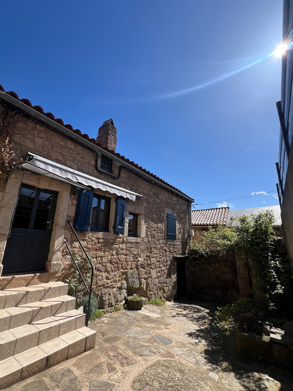 The exterior of the stone house is framed by a clear blue sky. A set of steps leads up to the entrance, accompanied by blue shutters and a chimney. Lush greenery surrounds the entryway, creating a serene outdoor space.
