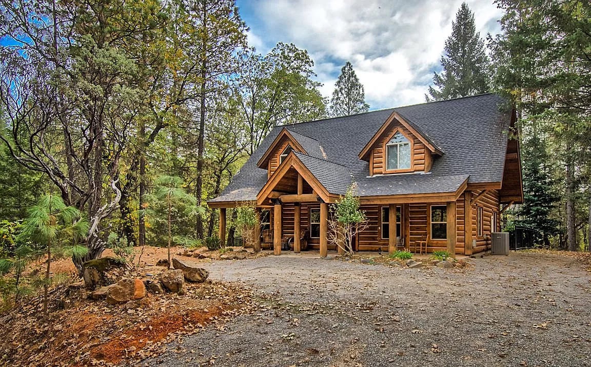 The log cabin features a spacious front porch under a gabled roof, surrounded by trees in a serene forest setting. Natural stone and wood elements are highlighted, with a gravel driveway leading up to the entrance.