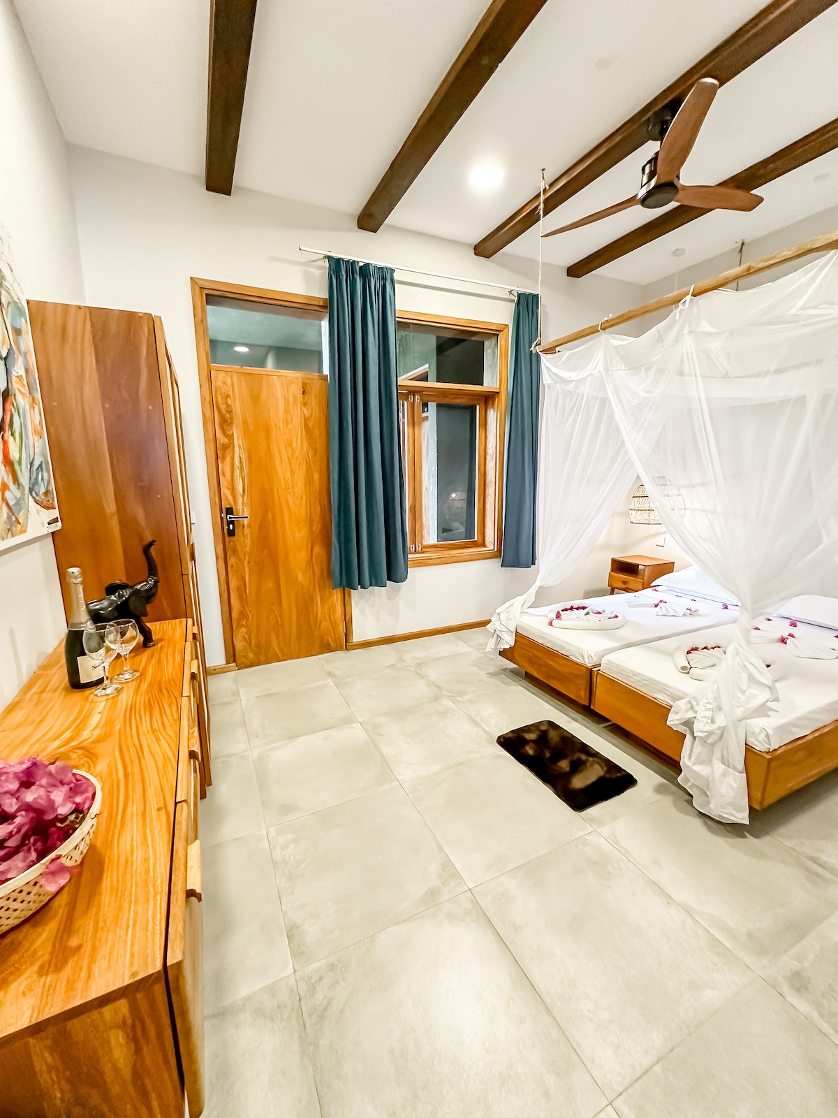 A cozy bedroom features a spacious bed adorned with decorative elements. Light-colored walls enhance the room's brightness, while dark curtains frame a large window. Natural materials are used in the wooden furniture, including a dresser and a side table with a welcoming gesture of champagne.