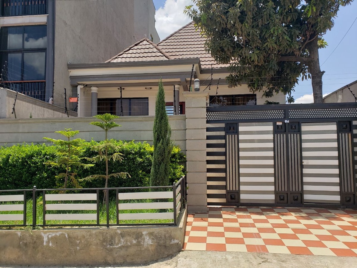 A residential villa is displayed, featuring a well-maintained garden with neatly trimmed hedges. The exterior showcases a symmetrical facade with a tiled roof and decorative columns. A gated entrance with horizontal slats provides a modern touch, while the driveway is accented by patterned tiles.