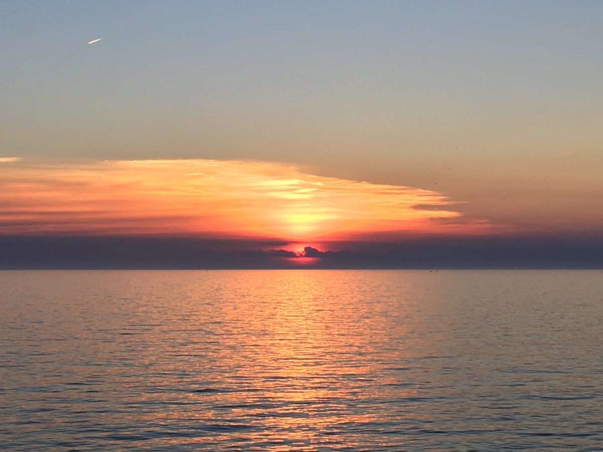 A serene sunset is captured over the calm waters of Lake Erie, with shades of orange, pink, and purple reflecting on the surface. The sky transitions from light to dark, creating a peaceful atmosphere at dusk.