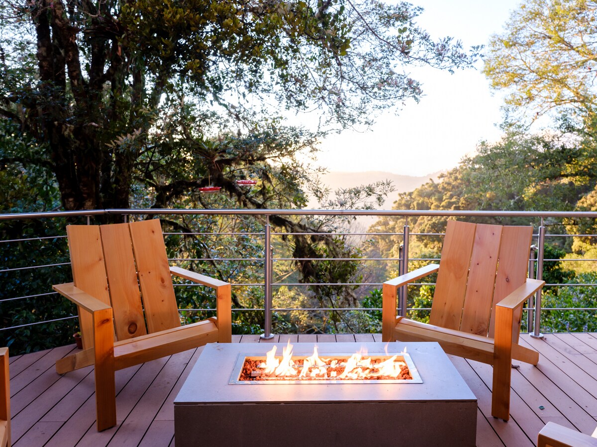 Two wooden adirondack chairs are positioned around a fire pit on a spacious deck. Lush trees surround the area, while distant hills are visible beyond the railing. The fire pit is lit, casting a warm glow, providing a cozy gathering spot amidst nature.