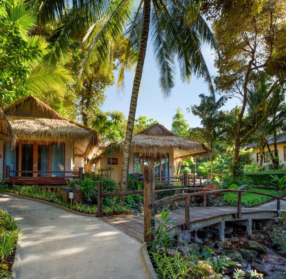 Two charming bungalows are nestled among lush greenery, featuring thatched roofs and wooden walkways. Palms sway gently in the breeze, providing shade. The pathway leads through well-maintained gardens, enhancing the natural beauty of the serene setting.