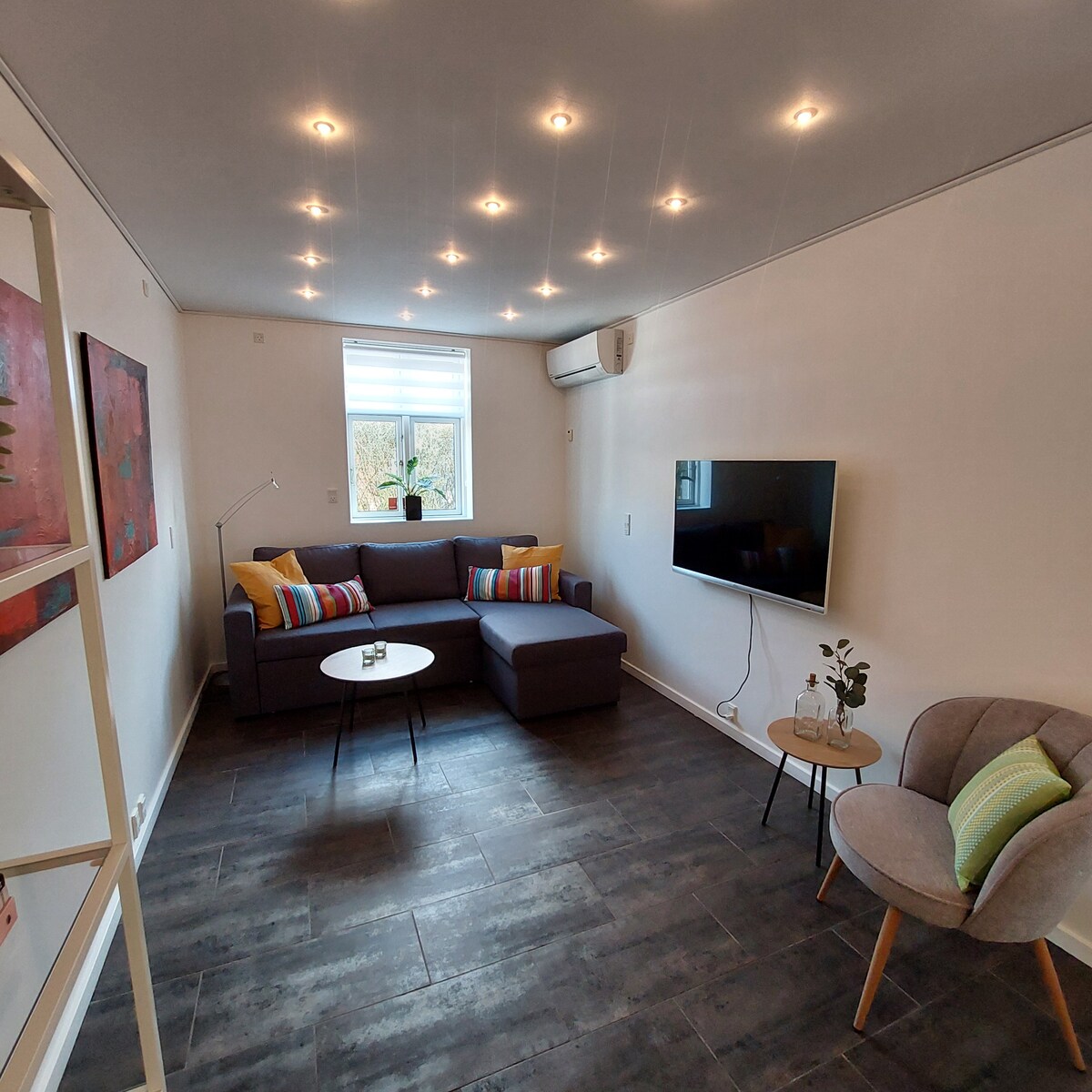 A comfortable living area features a sectional sofa adorned with colorful throw pillows. A small round table sits in front of the sofa, and a wall-mounted television is visible nearby. Natural light enters through a window, and recessed lighting enhances the space.