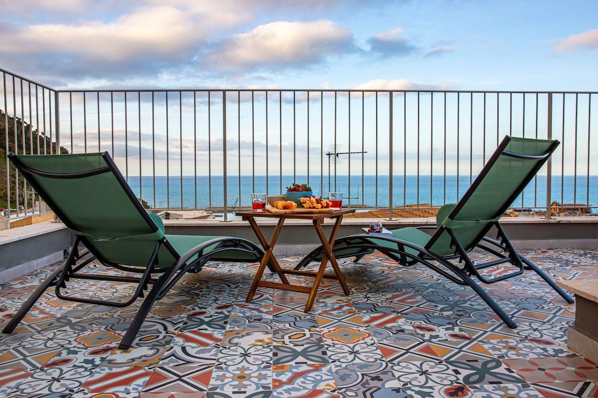 A terrace features two lounge chairs positioned near a wooden table. On the table, a selection of fruits and pastries is arranged. The background offers a view of the sea, with calm waters under a partly cloudy sky.