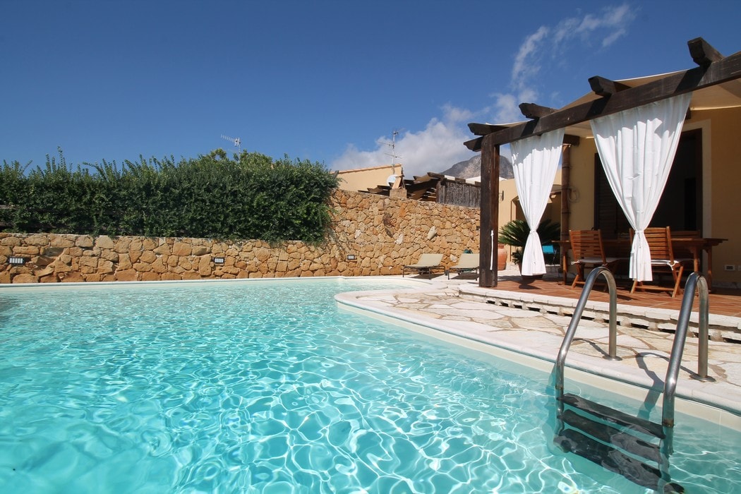 A private pool with clear blue water glistens under the sun, framed by elegant stone walls and lush greenery. A shaded patio area with wooden seating blends seamlessly into the outdoor space, offering a relaxing environment.