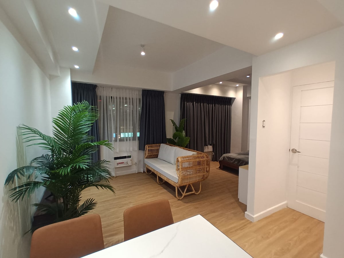 A spacious studio area features a rattan-style sofa and modern wooden flooring. Natural light filters through large windows, framed by dark curtains. A green potted plant adds a touch of nature, creating a relaxing environment. The layout includes a dining table visible in the foreground.