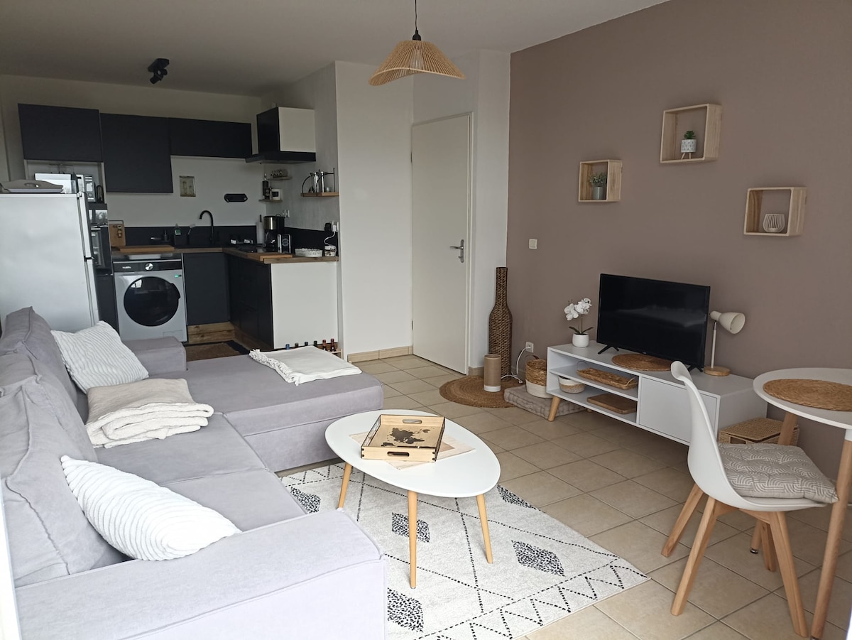The open-plan living area features a modern kitchen with new appliances and a cozy seating arrangement. A light grey sectional sofa sits invitingly across from a small dining table. Natural light fills the space, enhancing a relaxed atmosphere with neutral decor elements.