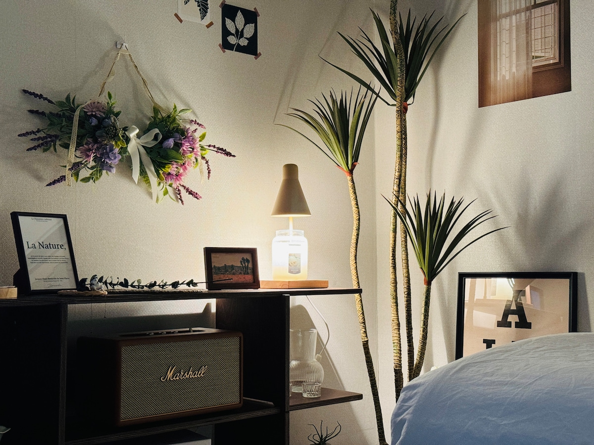 A corner of the room is showcased with decorative elements. A shelf displays framed art and a Marshall speaker, complemented by a bedside lamp. Green indoor plants add a touch of nature, while a floral arrangement hangs on the wall, enhancing the room's aesthetic appeal.